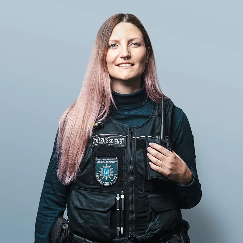 Eine leicht lächelnde langhaarige brünette Mitarbeiterin des Vollzugsdienstes in ihrer Uniform, die Hand legt sie an ihr in die Brusttasche eingestecktes Funkgerät